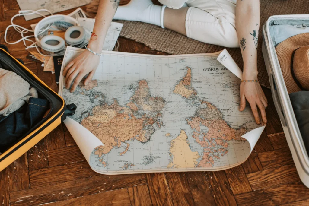 Overhead shot of a traveler sitting on a wooden floor unrolling a large world map between open suitcases while preparing for a trip