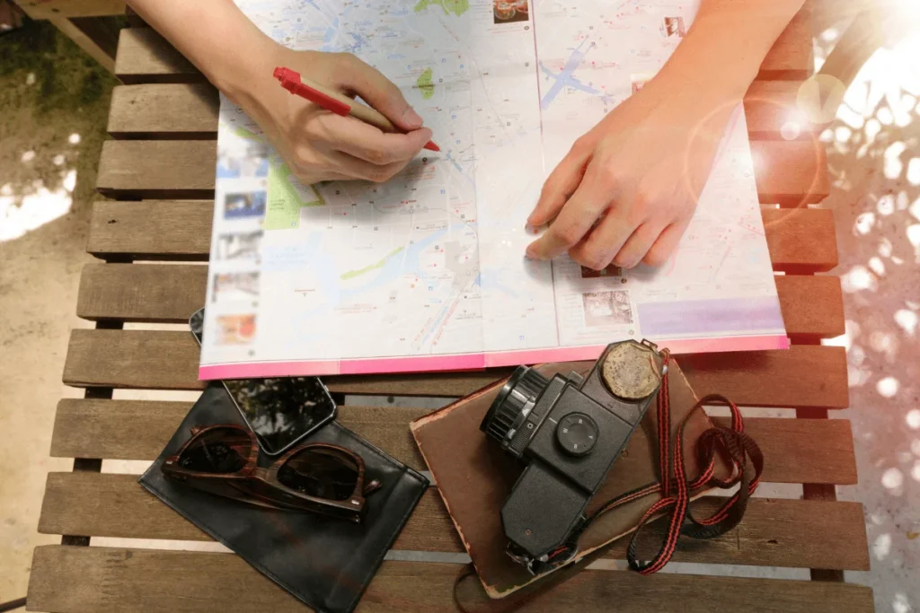 Top-down view of hands holding a red pen to mark locations on a folded paper travel map placed on a wooden slatted table alongside a vintage camera, leather case, and sunglasses.