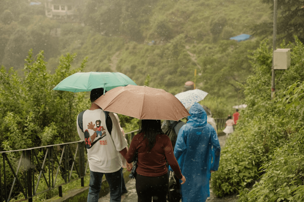 Travelers walking a hillside path in the rain with umbrellas and a blue rain poncho, surrounded by lush green vegetation